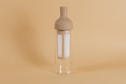 Tall glass container and white nylon mesh coffee filter, with mocha brown rubber wine bottle shaped top on an orange backdrop.