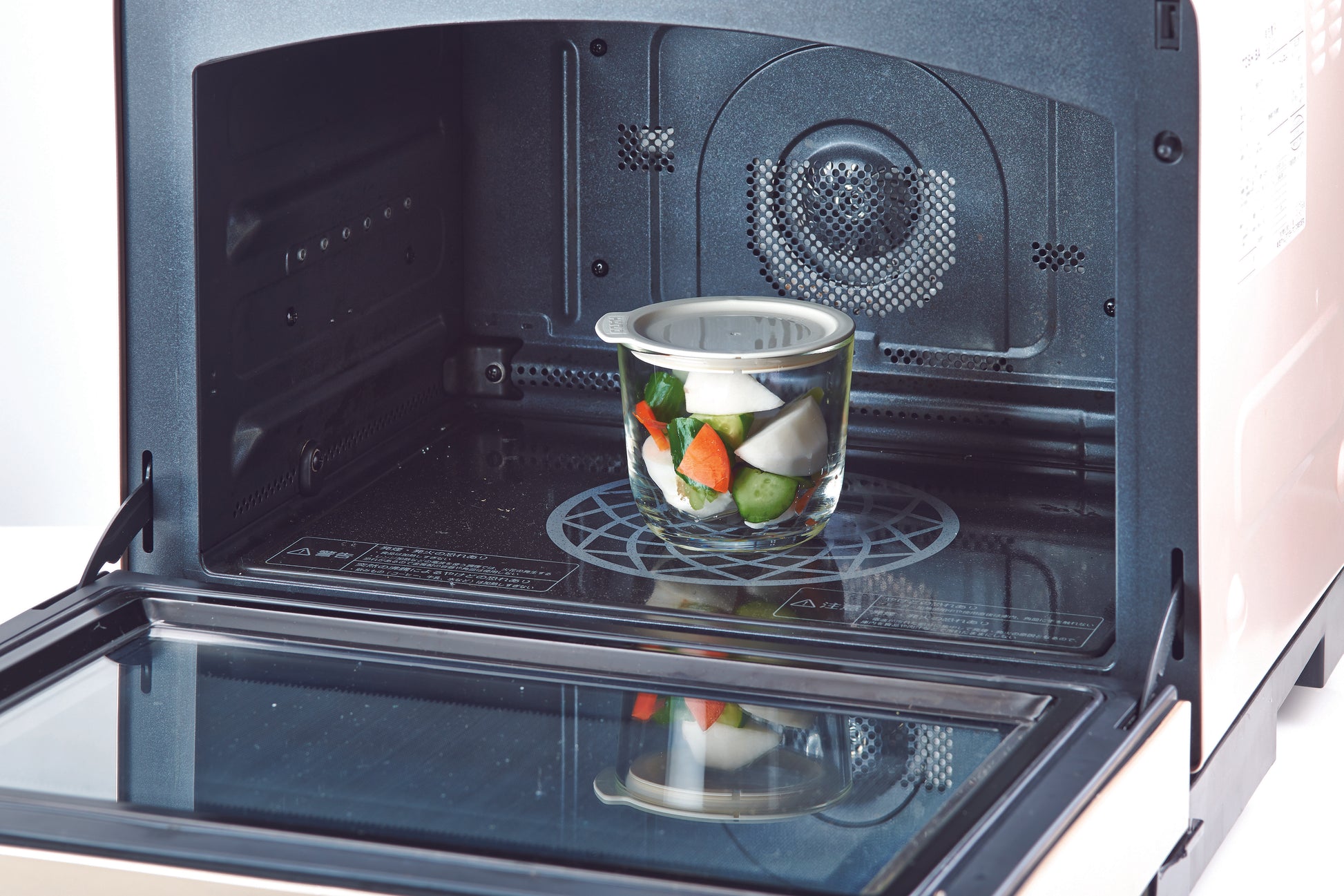 Clear glass salting bowl with white silicone lid with pickled vegetables inside sitting inside an open microwave with white exterior casing and black interior.