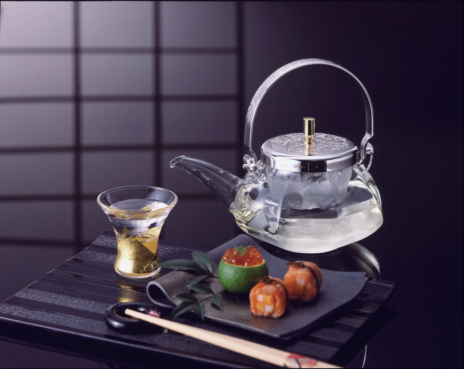 Elegant glass sake teapot with glass insert and metal lid and handle with decorative engraving set on a black reflective table with sushi and holding ice in its filter and sake in the pot. Tapered sake glass with gold trimmings and sake inside.
