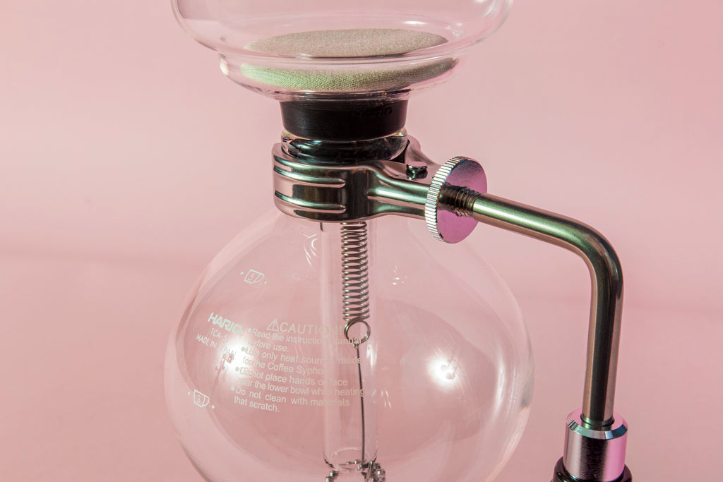 Close up of metal spring and cloth filter inside the glass serving and brewing chambers of a two piece glass coffee syphon attached to a metal and plastic arm with a metal adjustment pinion washer on a pink backdrop.