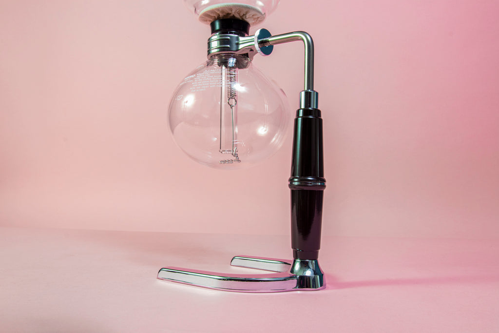 Close up of a two piece glass coffee syphon above an alcohol burner attached to a metal and plastic arm with a chrome base on a pink backdrop.