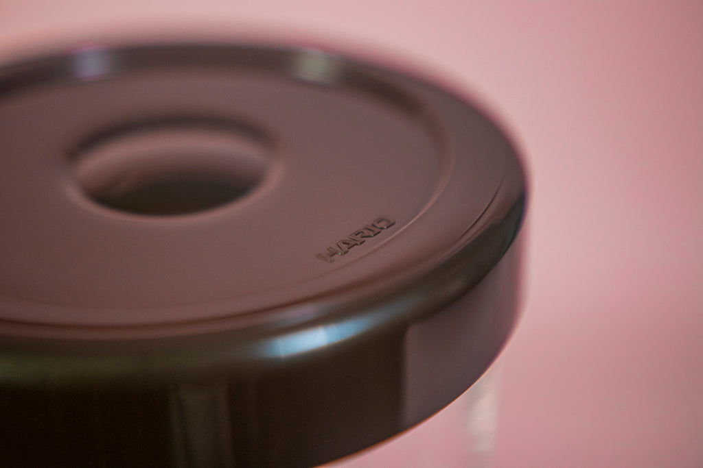 Close up of the Hario logo on a black plastic lid for the glass hopper of a two piece syphon coffee brewer on a pink background.