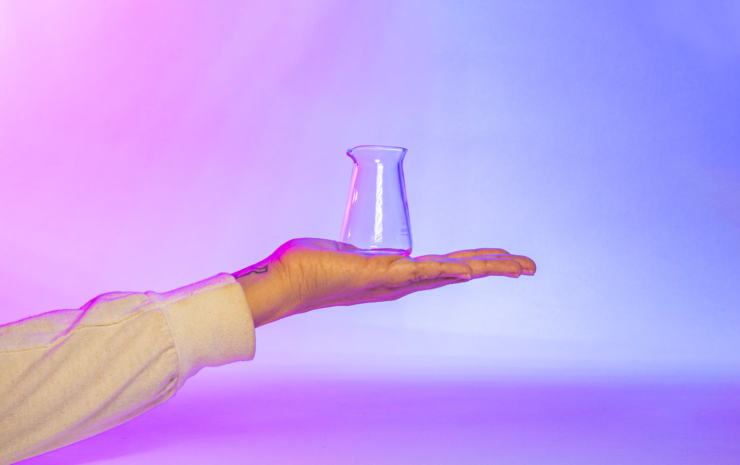 Tiny 50 millileter conical glass flask-shaped coffee pitcher sitting on open hand with white long sleeve shirt against a purple, pink, white, and blue background.