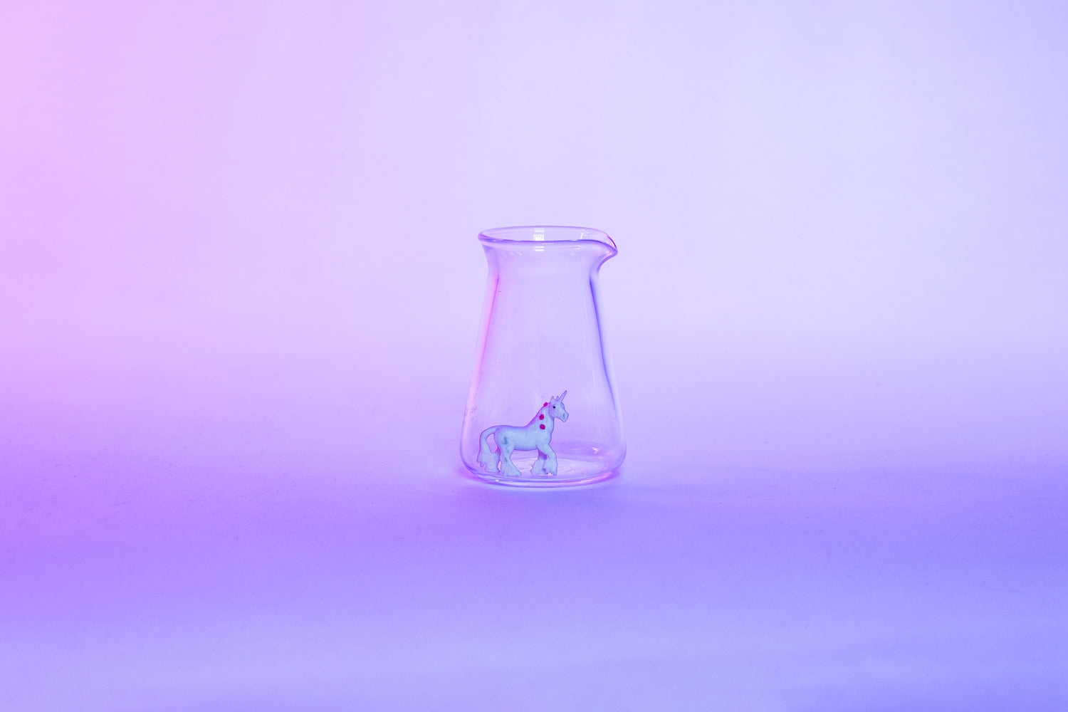 Conical glass flask-shaped coffee pitcher with small white unicorn toy inside and set against a light pink and purple background.