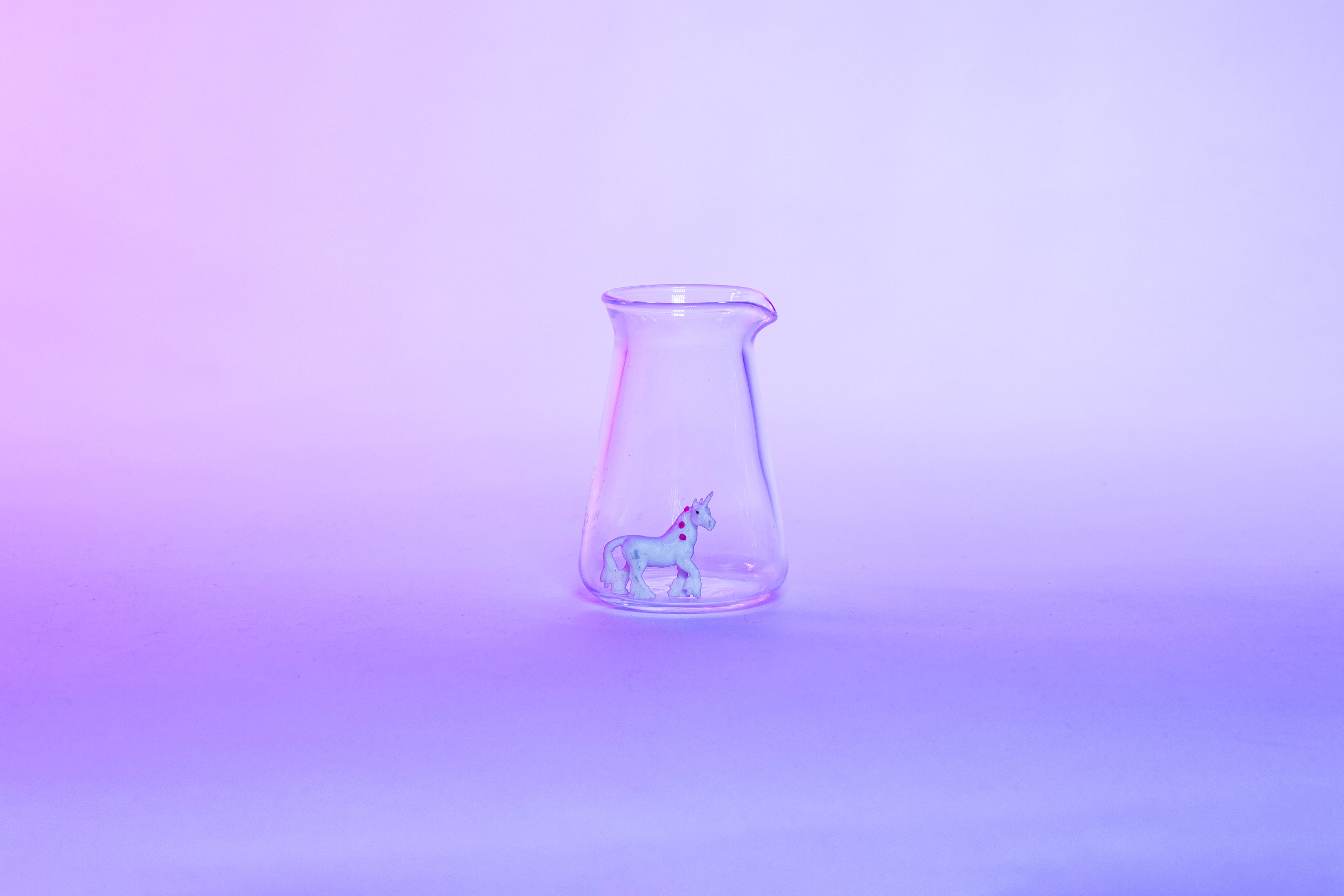 Conical glass flask-shaped coffee pitcher with small white unicorn toy inside and set against a light pink and purple background.