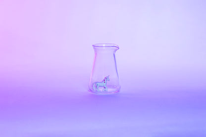 Conical glass flask-shaped coffee pitcher with small white unicorn toy inside and set against a light pink and purple background.