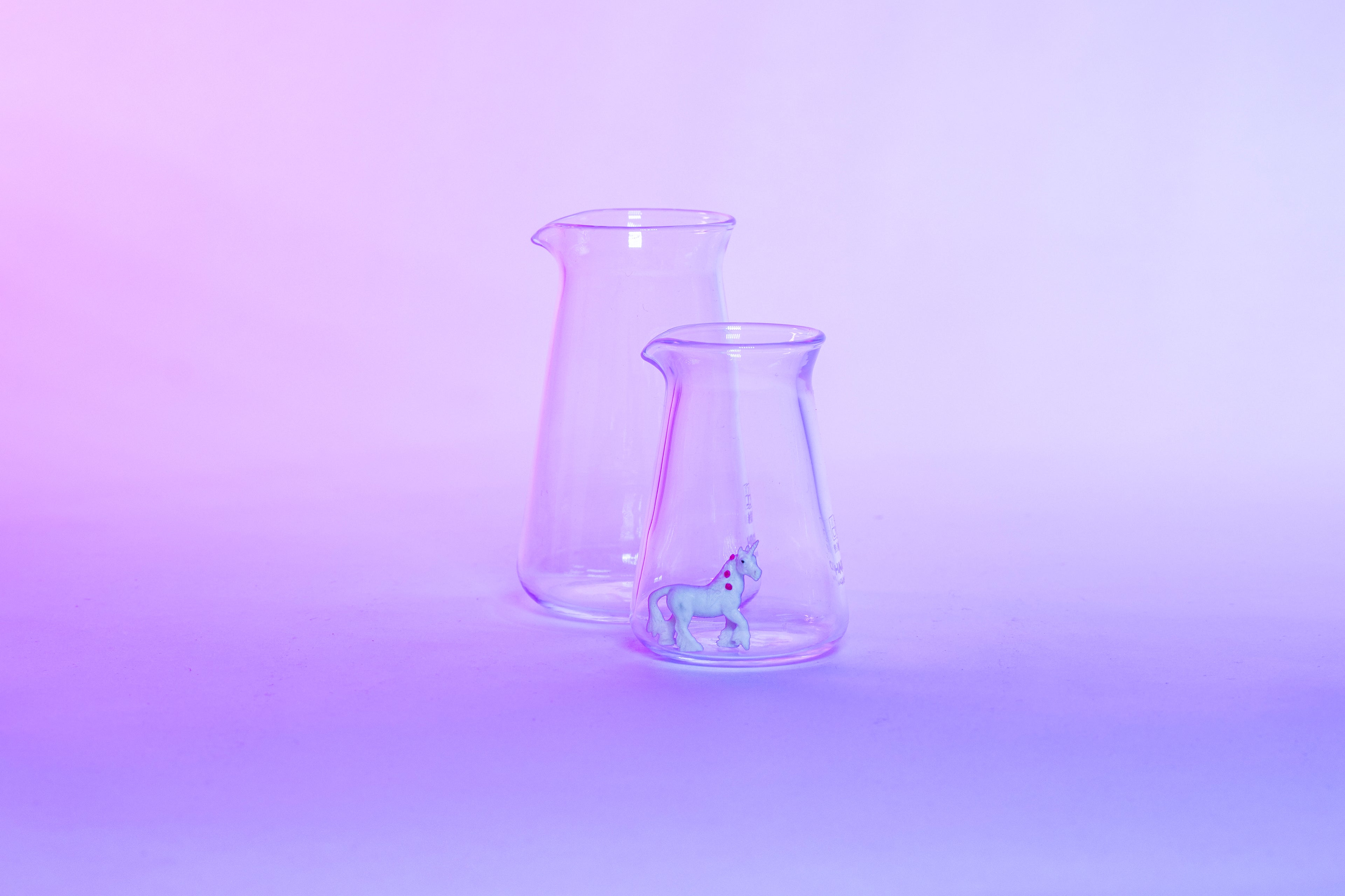 Two conical glass flask-shaped coffee pitchers, one big and one small, with a small white unicorn toy inside the small pitcher and set against a purple background.
