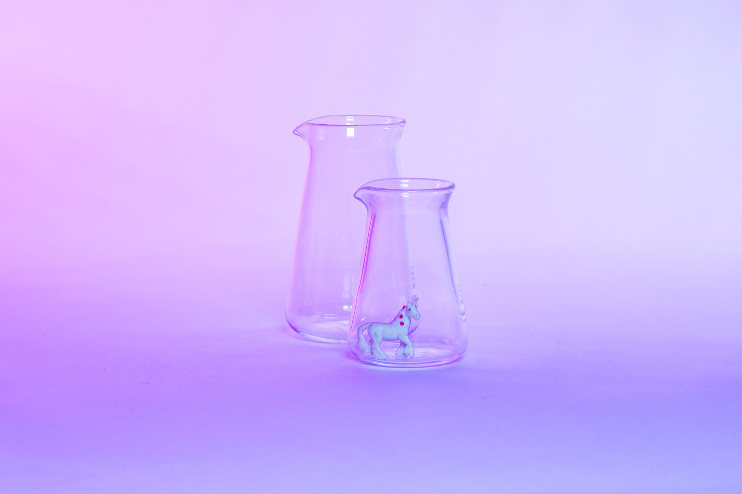 Two conical glass flask-shaped coffee pitchers, one big and one small, with a small white unicorn toy inside the small pitcher and set against a purple background.