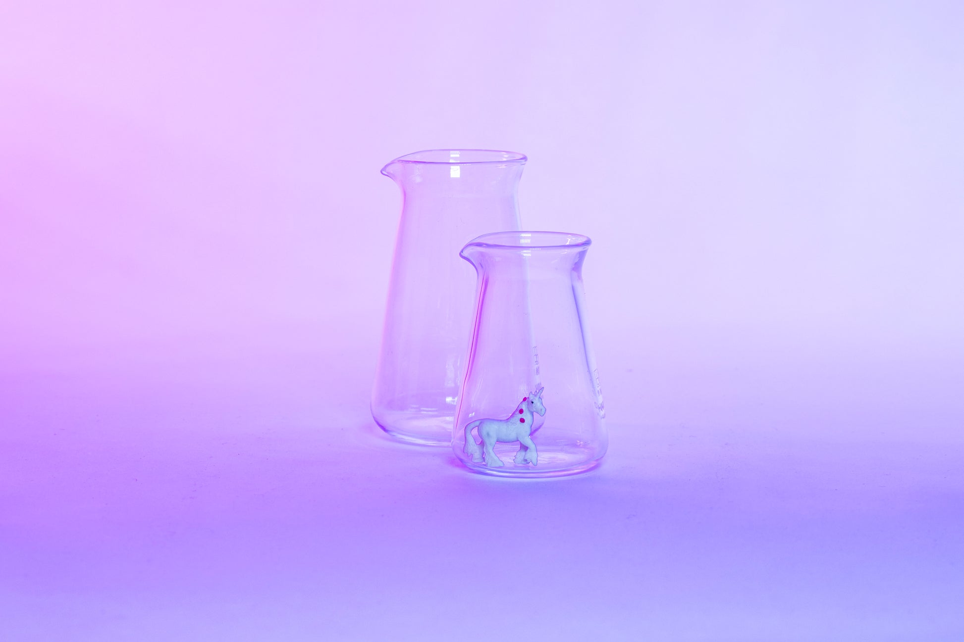 Two conical glass flask-shaped coffee pitchers, one big and one small, with a small white unicorn toy inside the small pitcher and set against a purple background.