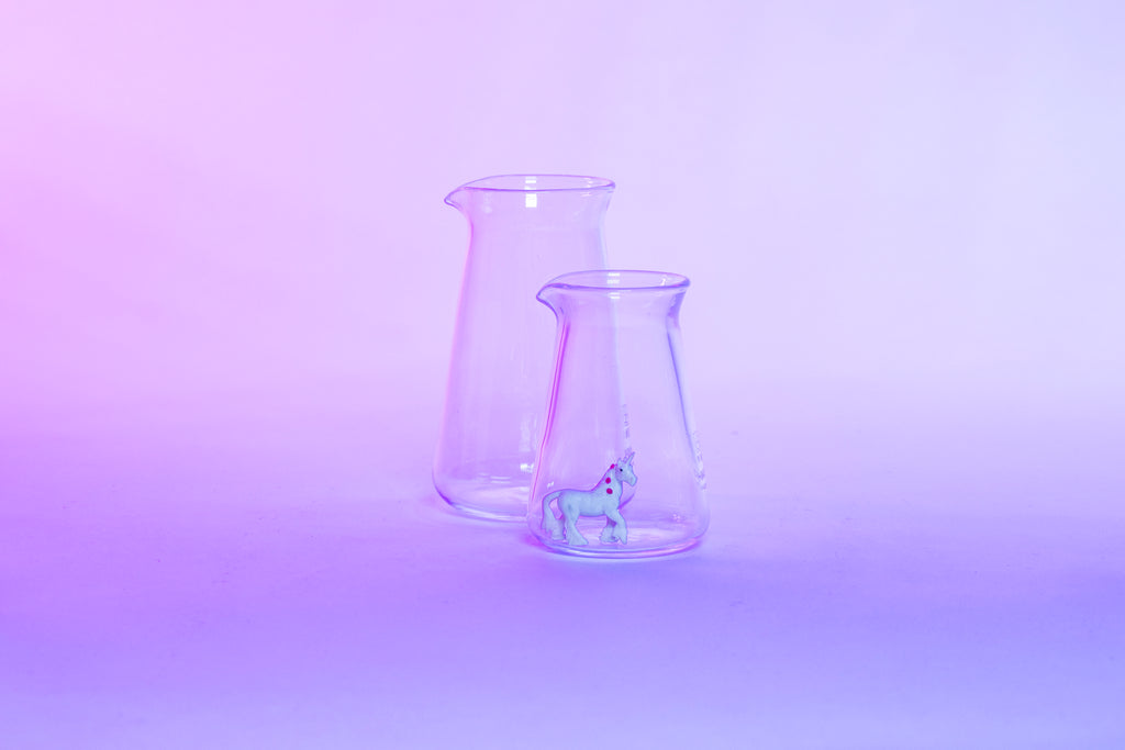 Two conical glass flask-shaped coffee pitchers, one big and one small, with a small white unicorn toy inside the small pitcher and set against a purple background.
