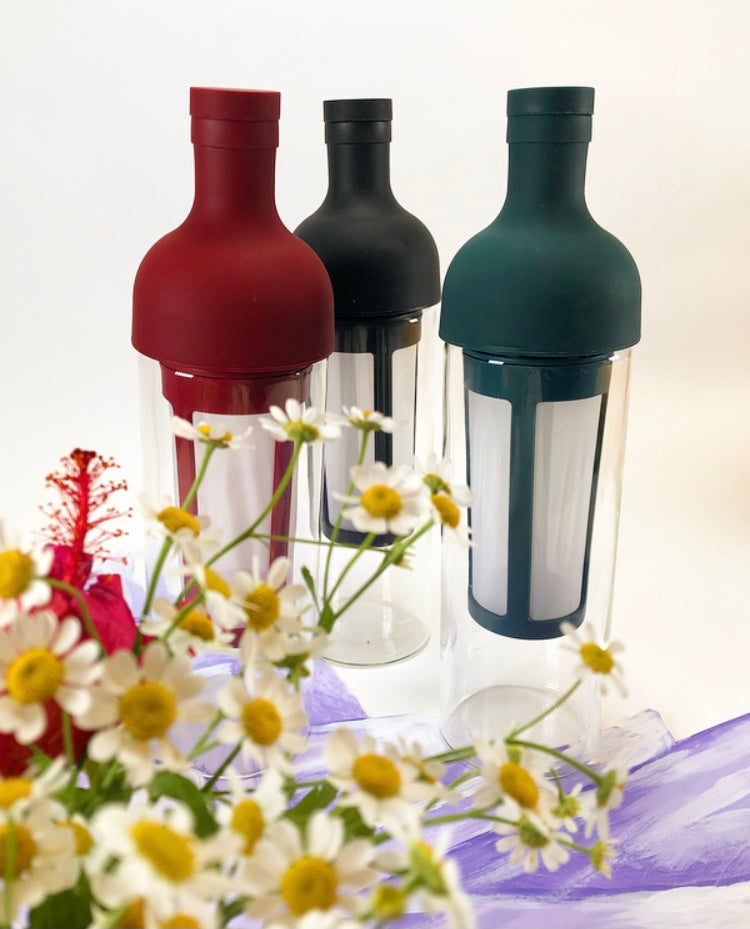 Three tall glass containers with white nylon mesh coffee filter and red, black, and teal rubber wine bottle shaped tops sitting behind white and yellow daisies and on top a purple sash.