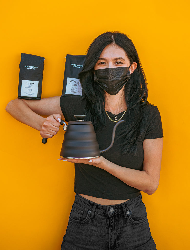 Model in all black with black mask and propping up two black bags of coffee on shoulder and elbow while holding a matte black metal gooseneck kettle and matching lid with plastic knob and black plastic covered handle against a yellow background.