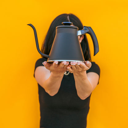 Model holding a matte black kettle with gooseneck spout, sloped black plastic handle and flat, black plastic lid knob with two open palms and set against a yellow background.