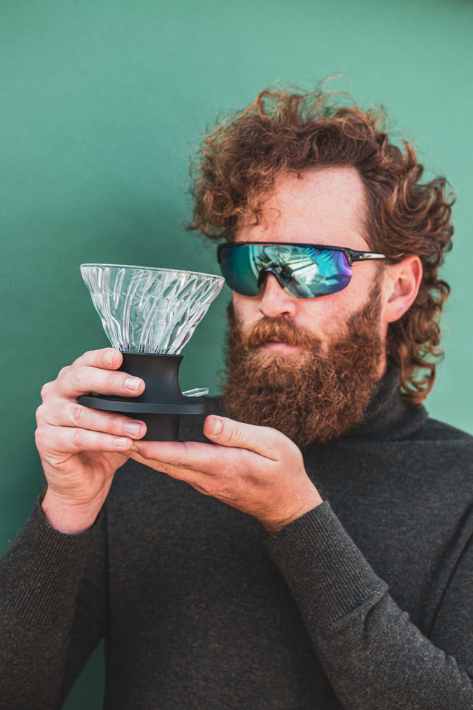 Man with beard and sunglasses holding a cone-shaped glass dripper seated in a black plastic silicone base with clear plastic push switch on a blue-green background.