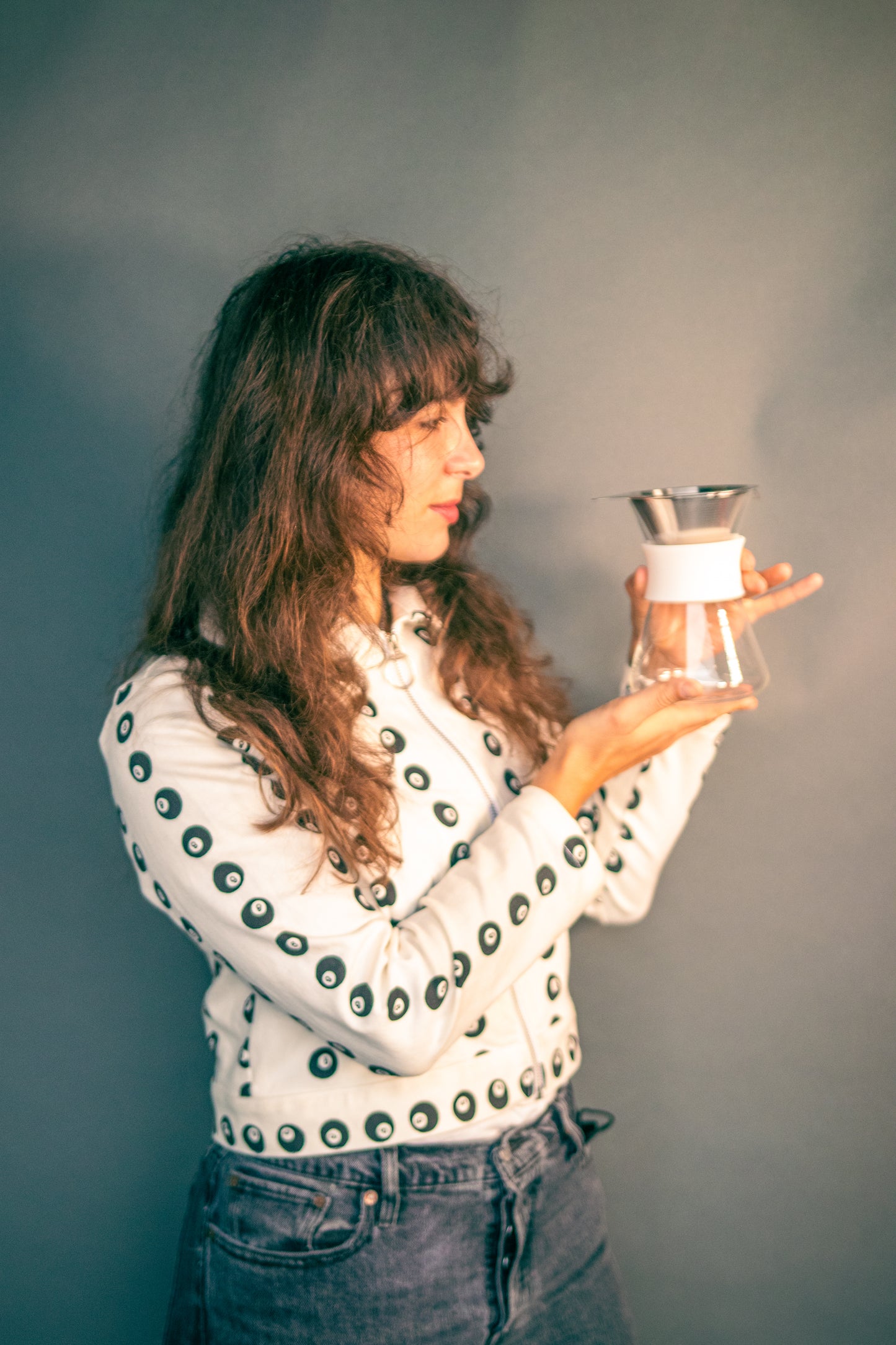 Woman holding a clear glass server with white silicone collar and stainless steel mesh filter against a grey background.