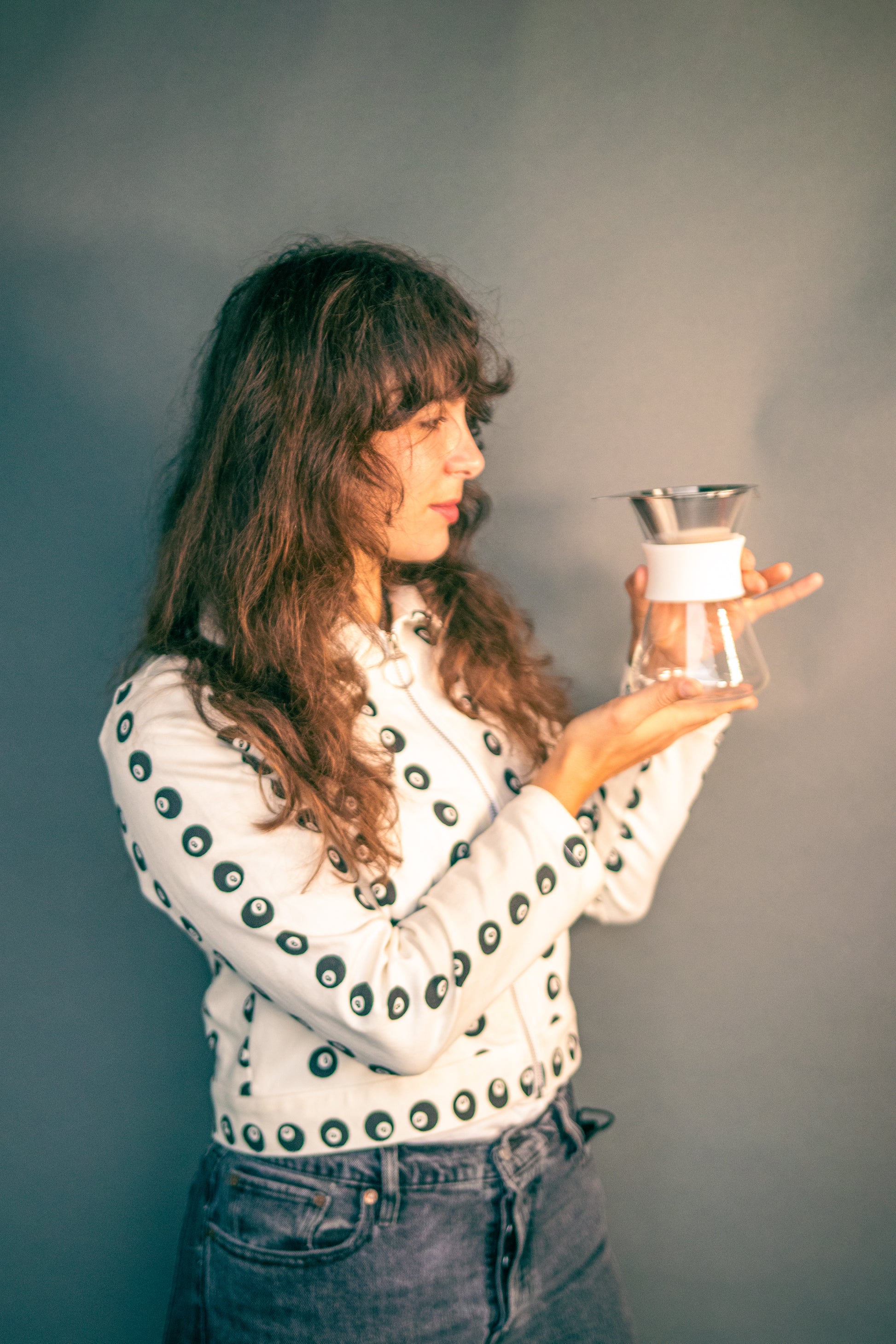 Woman holding a clear glass server with white silicone collar and stainless steel mesh filter against a grey background.