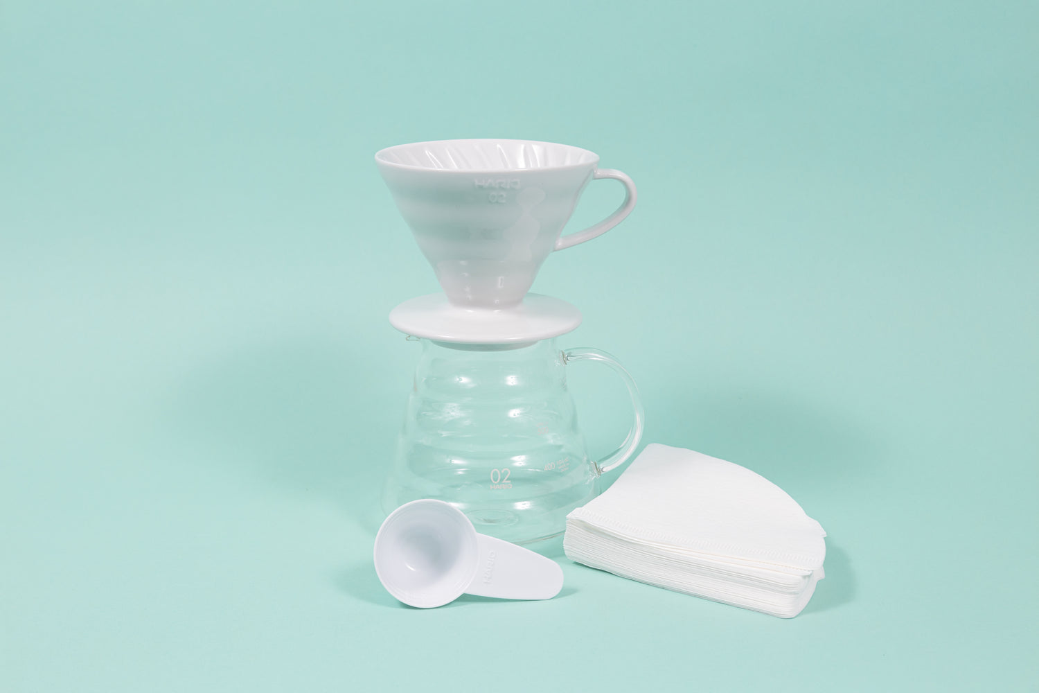 White ceramic conical coffee dripper with handle on top of an all glass coffee server with handle and plastic scoop and filter stack.