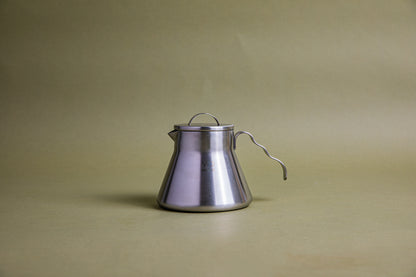 Stainless steel beaker-shaped coffee server with stainless steel lid, u-shaped lid handle, and open wavy stainless steel handle. This server is designed for camping and is set against an earth-toned background.