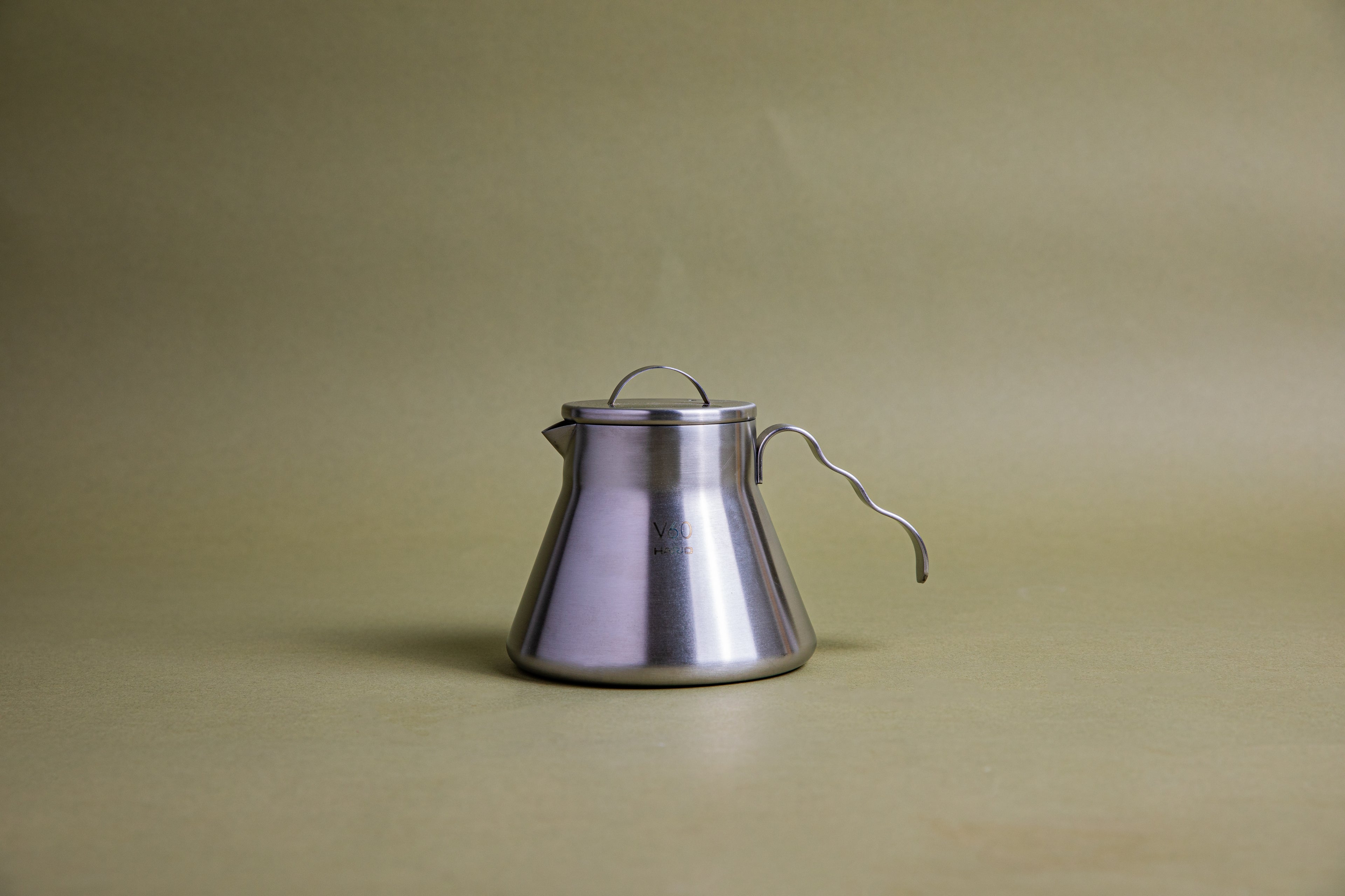 Stainless steel beaker-shaped coffee server with stainless steel lid, u-shaped lid handle, and open wavy stainless steel handle. This server is designed for camping and is set against an earth-toned background.