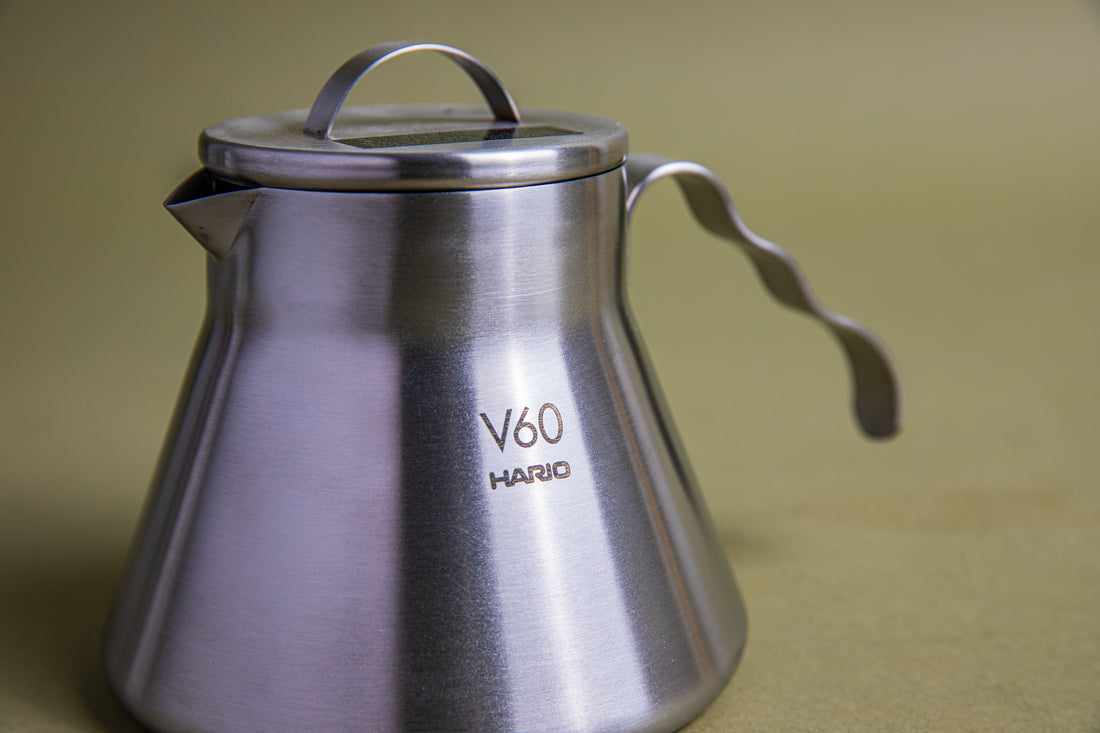 Close up of a stainless steel beaker-shaped coffee server with stainless steel lid, u-shaped lid handle, and open wavy stainless steel handle. This server is designed for camping and is set against an earth-toned background.