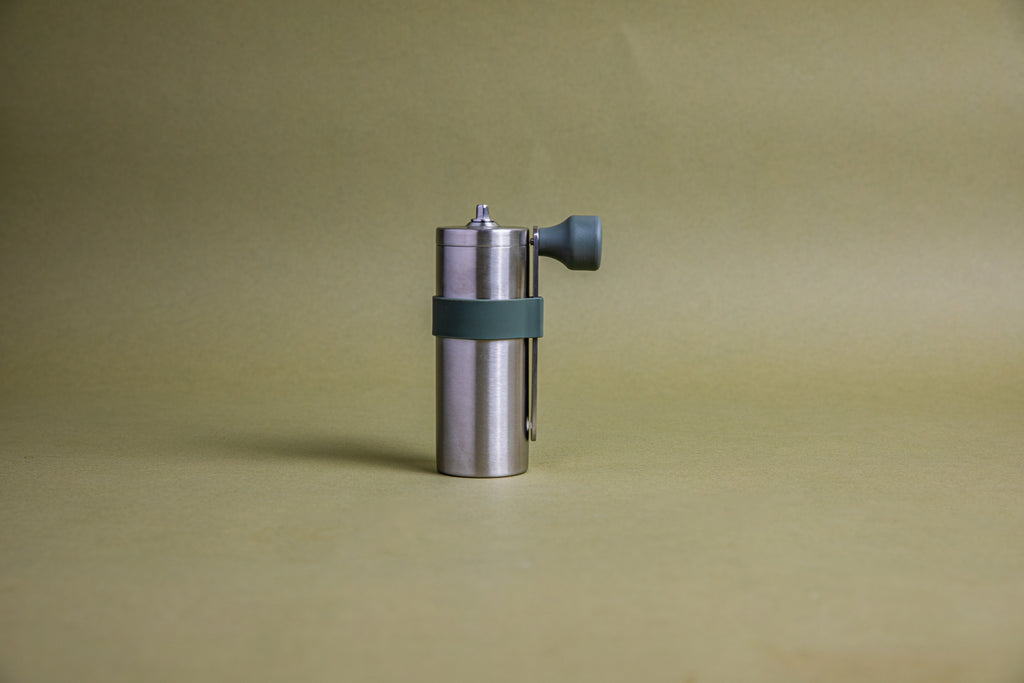 Stainless steel cylindrically shaped coffee mill cannister with forest green silicone band holding stainless steel handle with plastic knob. Set against an earth-tone background.
