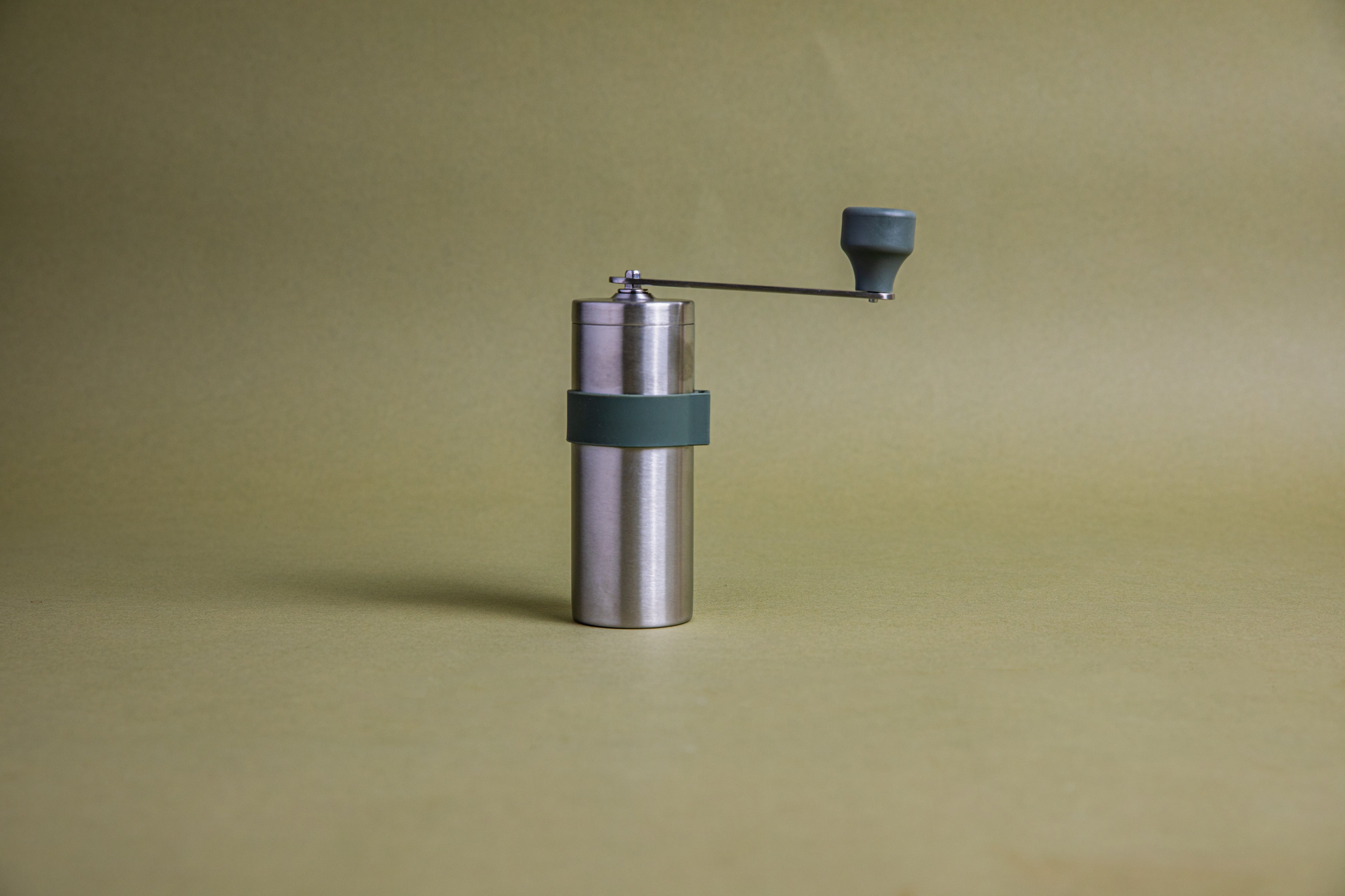 Stainless steel cylindrically shaped coffee mill cannister with forest green silicone band stainless steel handle with plastic knob. Set against an earth-tone background.