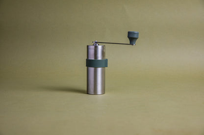 Stainless steel cylindrically shaped coffee mill cannister with forest green silicone band stainless steel handle with plastic knob. Set against an earth-tone background.