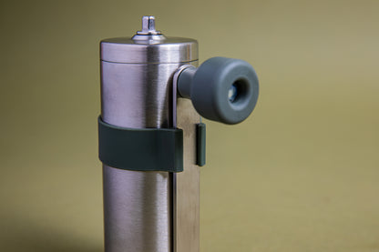 Close up of a stainless steel cylindrically shaped coffee mill cannister with forest green silicone band holding stainless steel handle with plastic knob. Set against an earth-tone background.