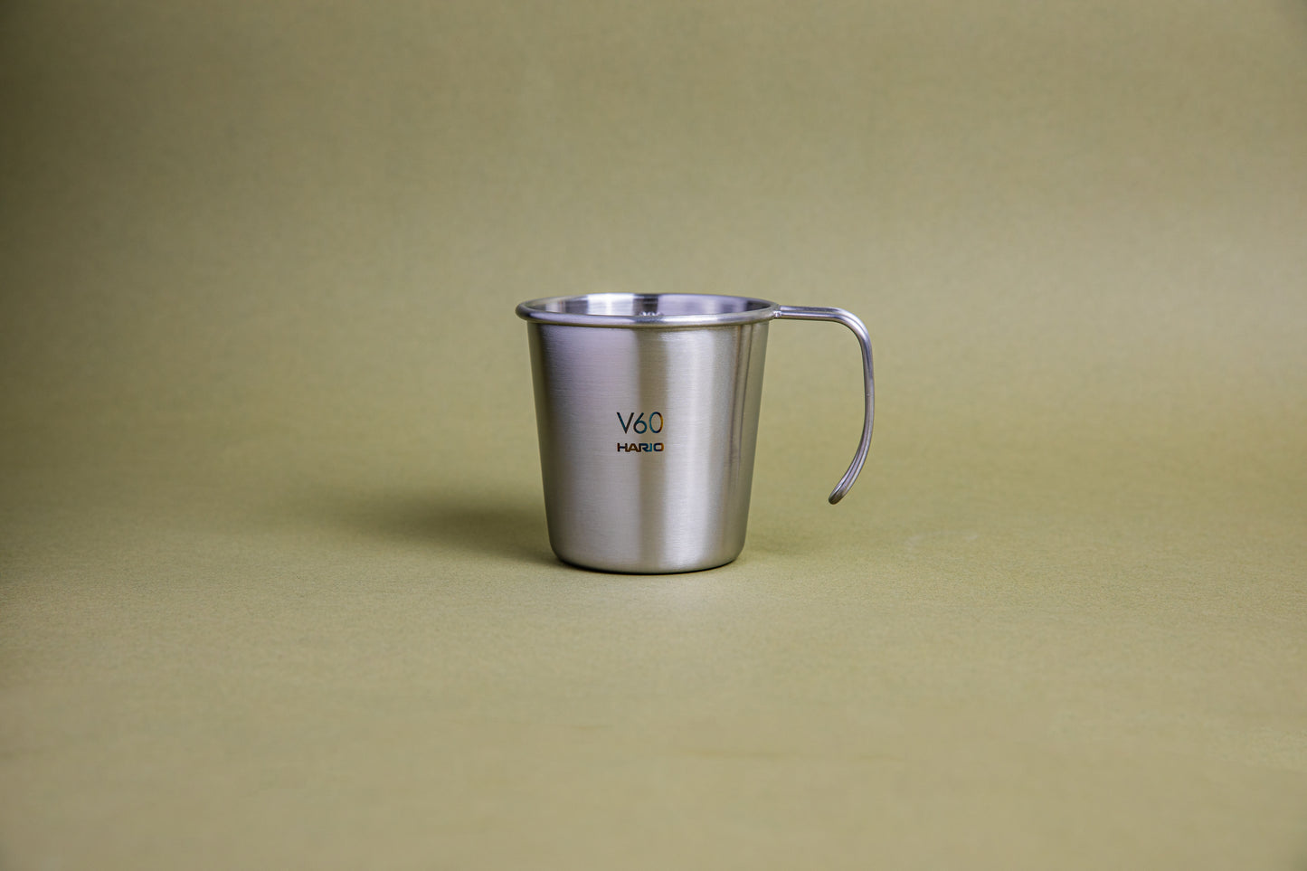 Sleek hairline silver camping mug with rounded lip and handle, etched Hario logo and V60 graphic, and set against a earth-tone background.
