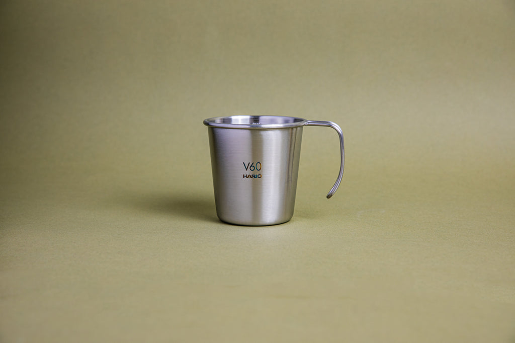 Sleek hairline silver camping mug with rounded lip and handle, etched Hario logo and V60 graphic, and set against a earth-tone background.