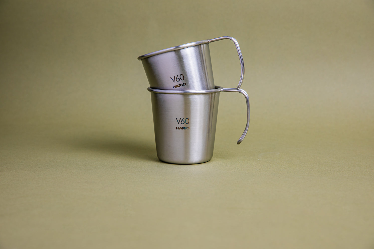 Two sleek hairline silver camping mugs with rounded lips and open handles, etched Hario logos and V60 graphics, stacked in each other, and set against a earth-tone background.