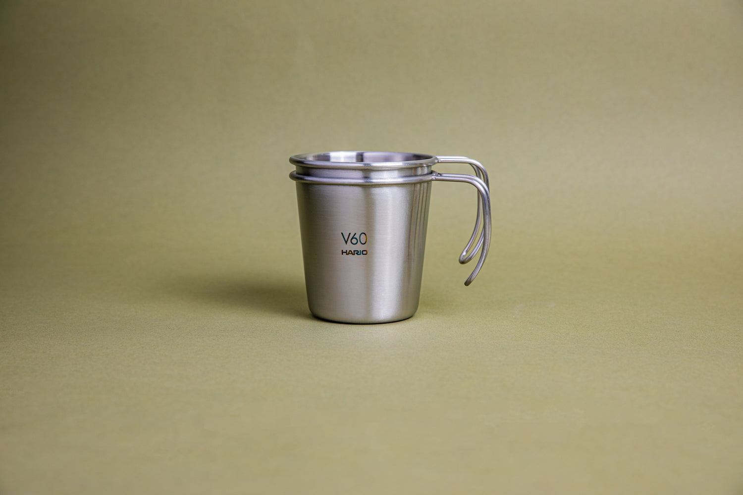 A sleek hairline silver camping mug with rounded lip and handle, etched Hario logo and V60 graphic, and set against a earth-tone background with another stacking mug stacked inside..