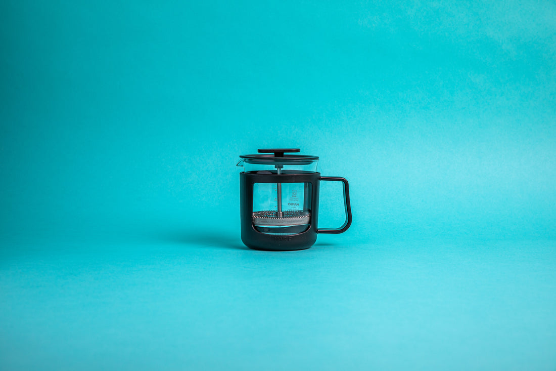 Coffee press featuring a glass pitcher with fluted spout, stainless steel mesh filter with pole, and a black plastic lid, knob, holder, and handle. Set against a blue background.