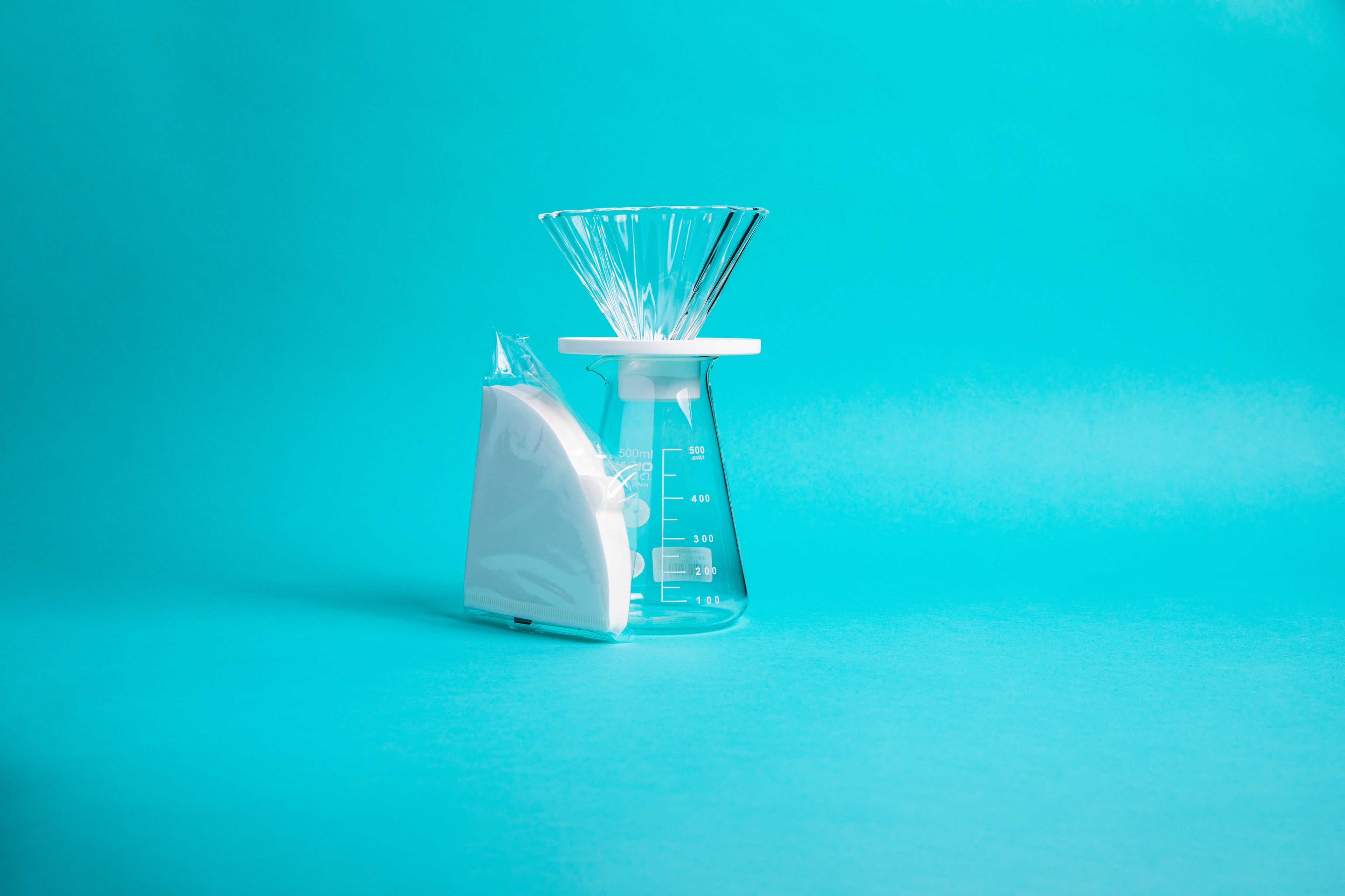Fluted, flower-shaped glass cone with white plastic holder seated atop a tapered glass beaker with white millileter measurement marks and fluted spout, next to a pack of white cone-shaped filters in a clear plastic wrapper, and set against a blue background.