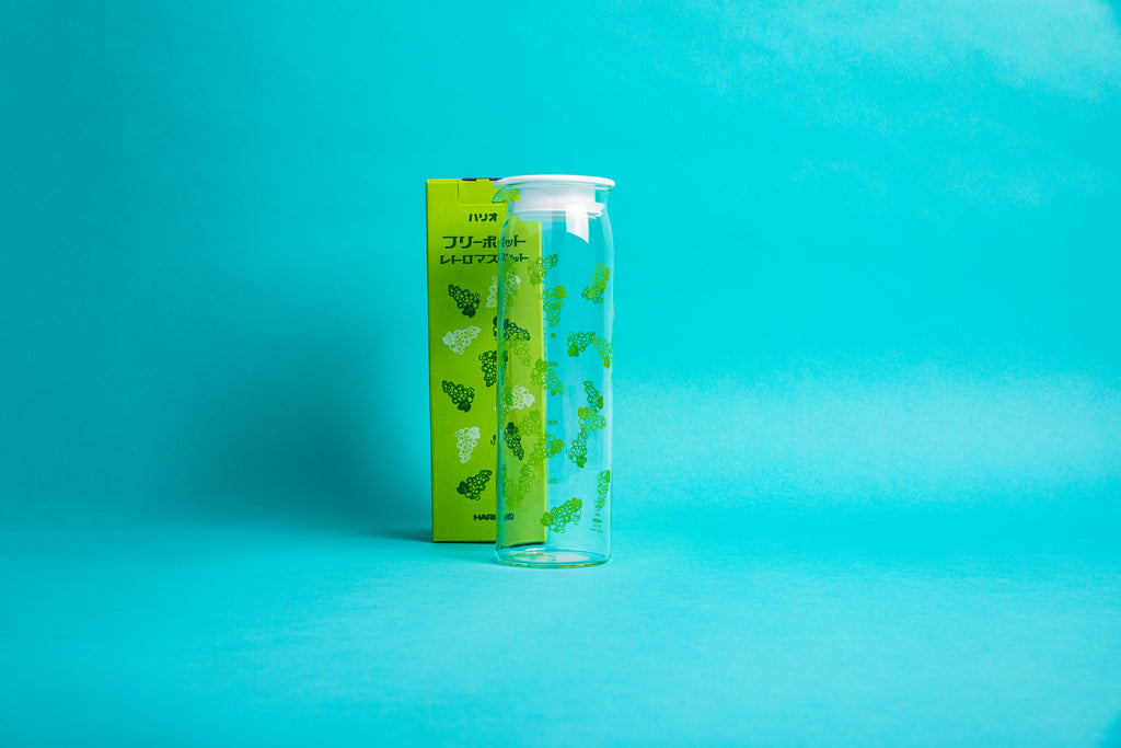 Tall cylindrically shaped glass pot with fluted spout, green grape pattern, and white plastic lid in front of its yellow packaging box with red and white grape pattern and set against a blue background.