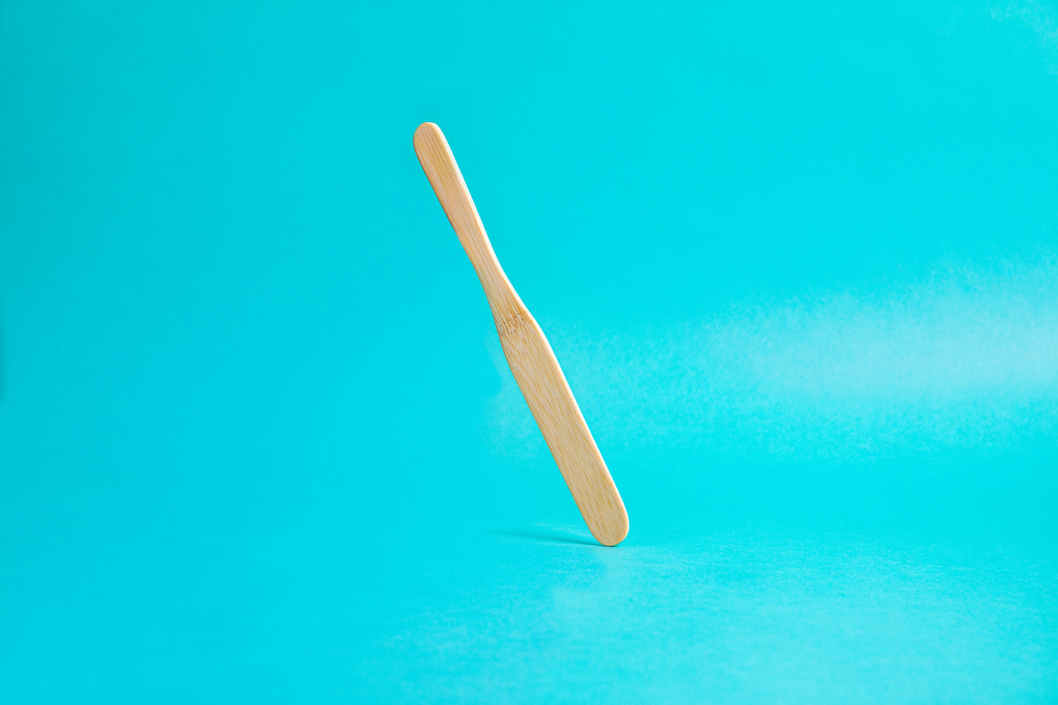 Flat, smooth, bamboo stirring stick with rounded edges, tapered between the handle and stirring portion, and set against a blue background. This item is remniscent in shape to a tongue depressor and works great for evenly stirring coffee grounds.