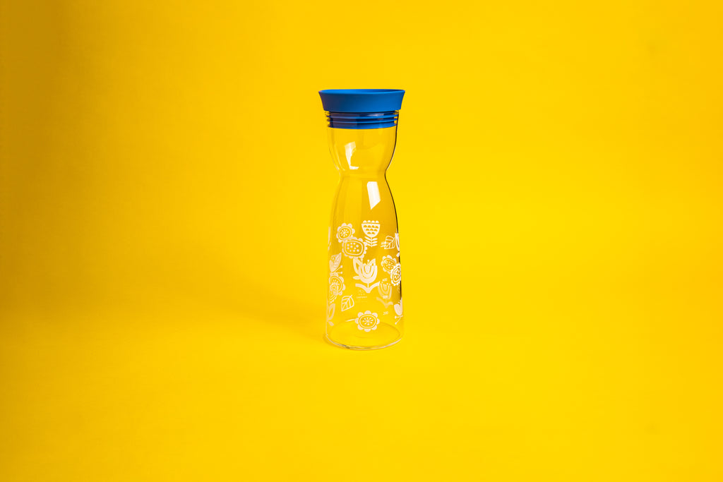 Hourglass shaped glass with white flower etching. Topped with a blue silicone cap. Set on a yellow background.