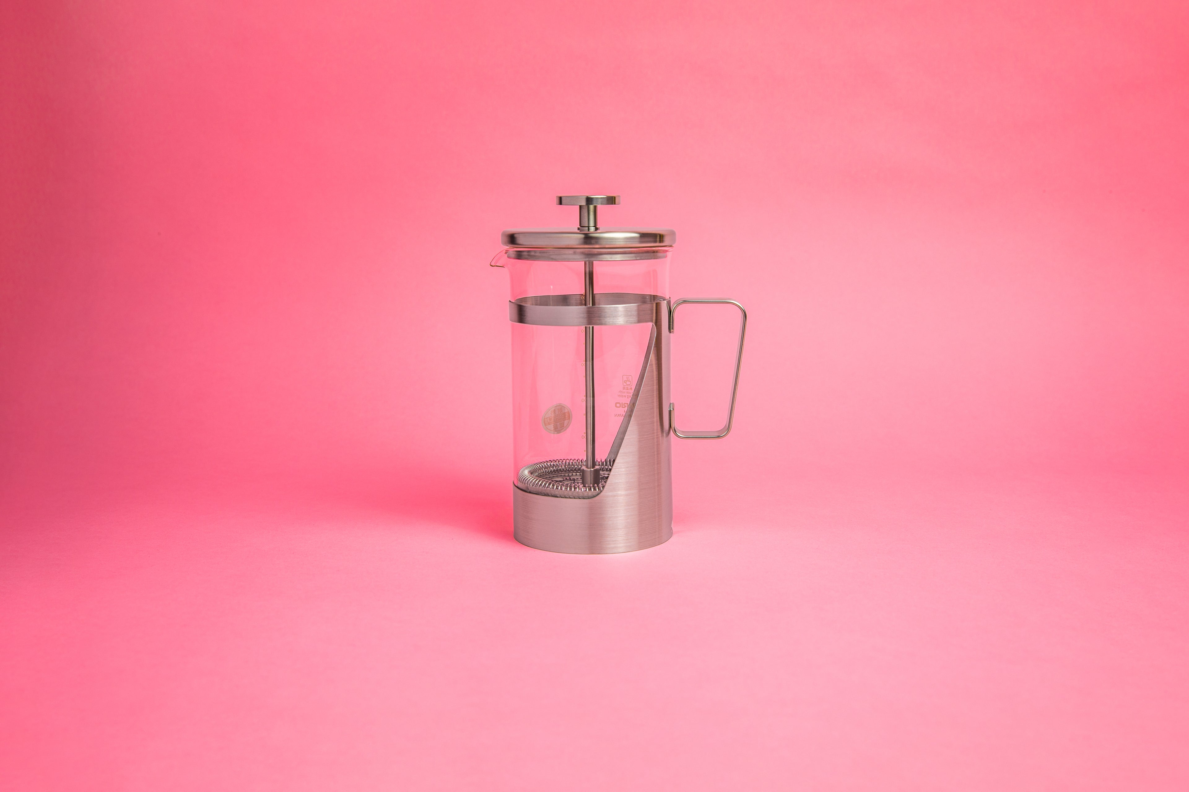 Coffee press featuring a glass pitcher with fluted spout, stainless steel mesh filter with pole, and a stainless steel lid, knob, holder, and handle. Set against a pink background.