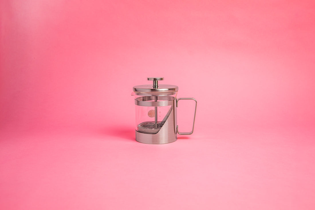 Coffee press featuring a glass pitcher with fluted spout, stainless steel mesh filter with pole, and a stainless steel lid, knob, holder, and handle. Set against a pink background.