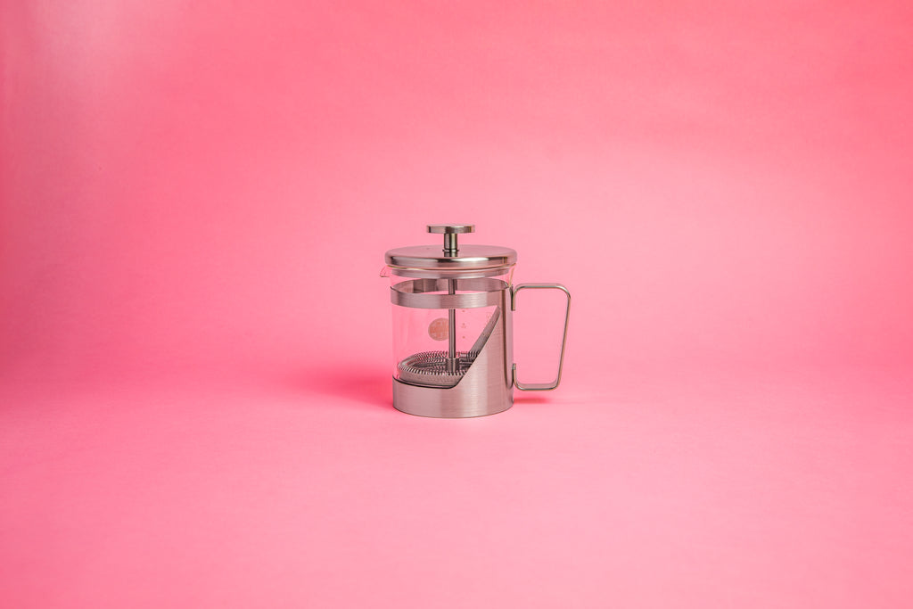 Coffee press featuring a glass pitcher with fluted spout, stainless steel mesh filter with pole, and a stainless steel lid, knob, holder, and handle. Set against a pink background.