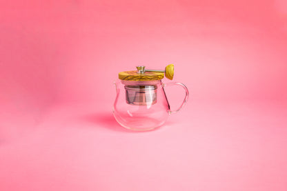 Glass teapot with glass handle. Olive wood lid and knob. Steel rod with a steel tea filter basket.  set on pink gradient background