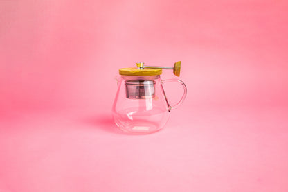 Glass teapot with glass handle. Olive wood lid and knob. Steel rod with a steel tea filter basket.  set on pink gradient background