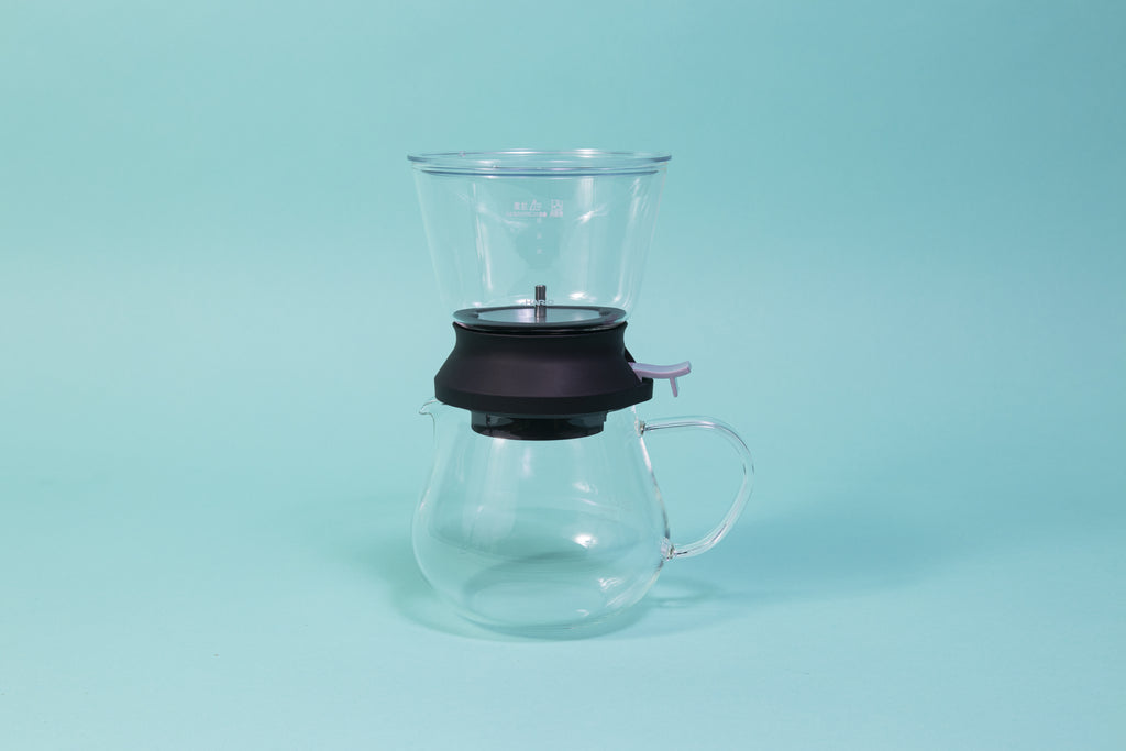 Cylindrical glass tea brewer and metal mesh strainer with clear plastic lid atop a black rubber base with plastic lever sitting on all glass bulbous server