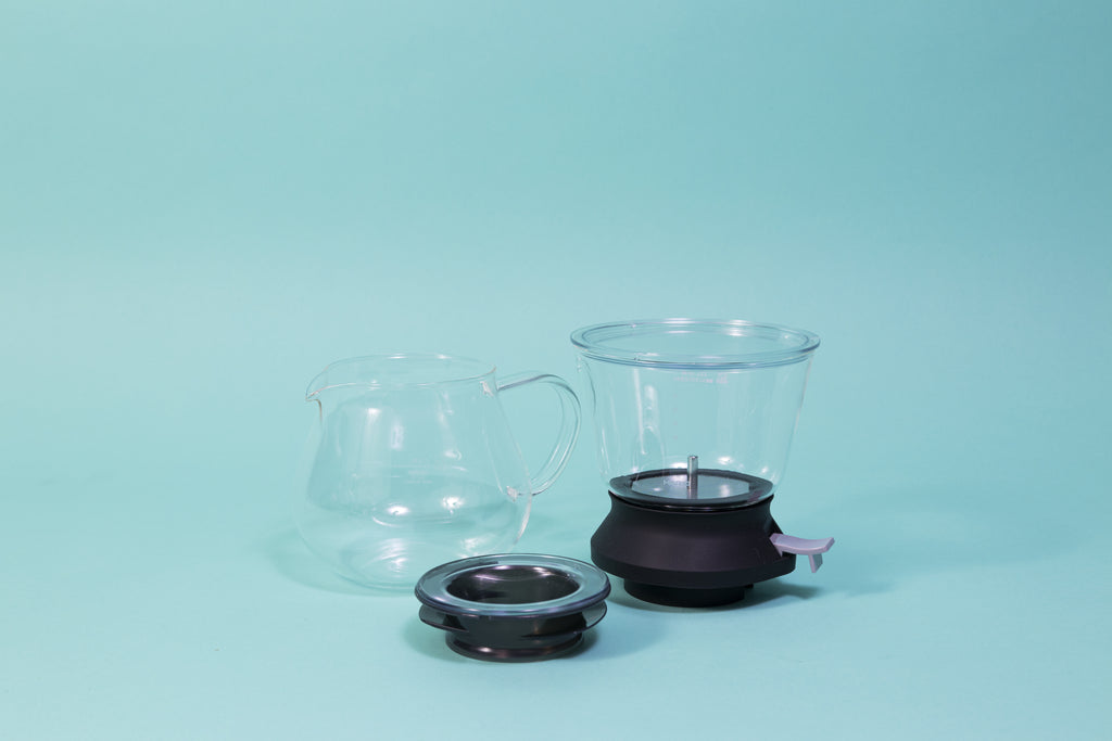Cylindrical glass tea brewer and metal mesh strainer with clear plastic lid atop a black rubber base with plastic lever sitting next to all glass bulbous server and transparent black lid