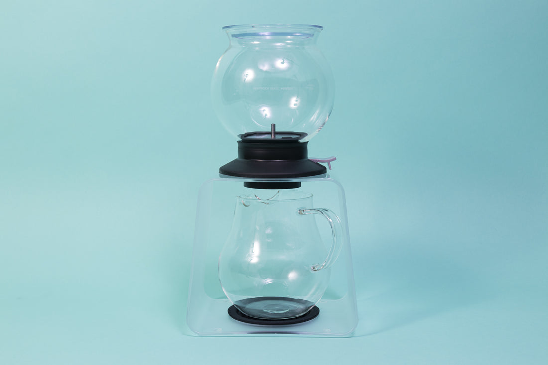 Globulous glass tear brewer and metal mesh filter with clear plastic lid atop a black rubber base with plastic lever switch on a clear acrylic stand above an all glass bulbous server with handle