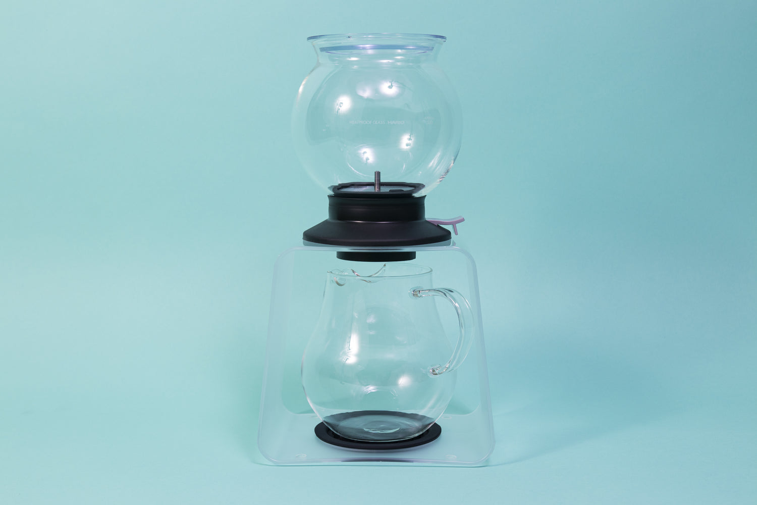 Globulous glass tear brewer and metal mesh filter with clear plastic lid atop a black rubber base with plastic lever switch on a clear acrylic stand above an all glass bulbous server with handle
