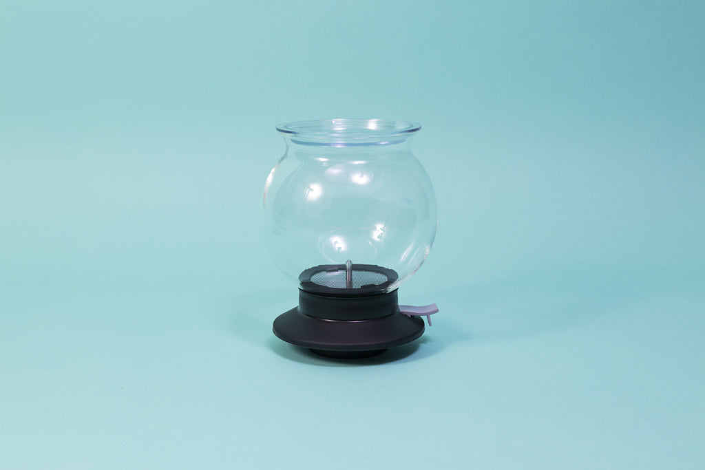 Globulous glass tear brewer and metal mesh filter with clear plastic lid atop a black rubber base with plastic lever switch 