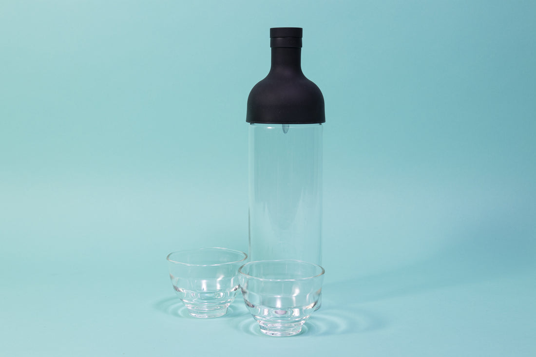 Tall glass container with black rubber wine bottle shaped top on a blue backdrop with two honey-comb shaped short glass tea cups.