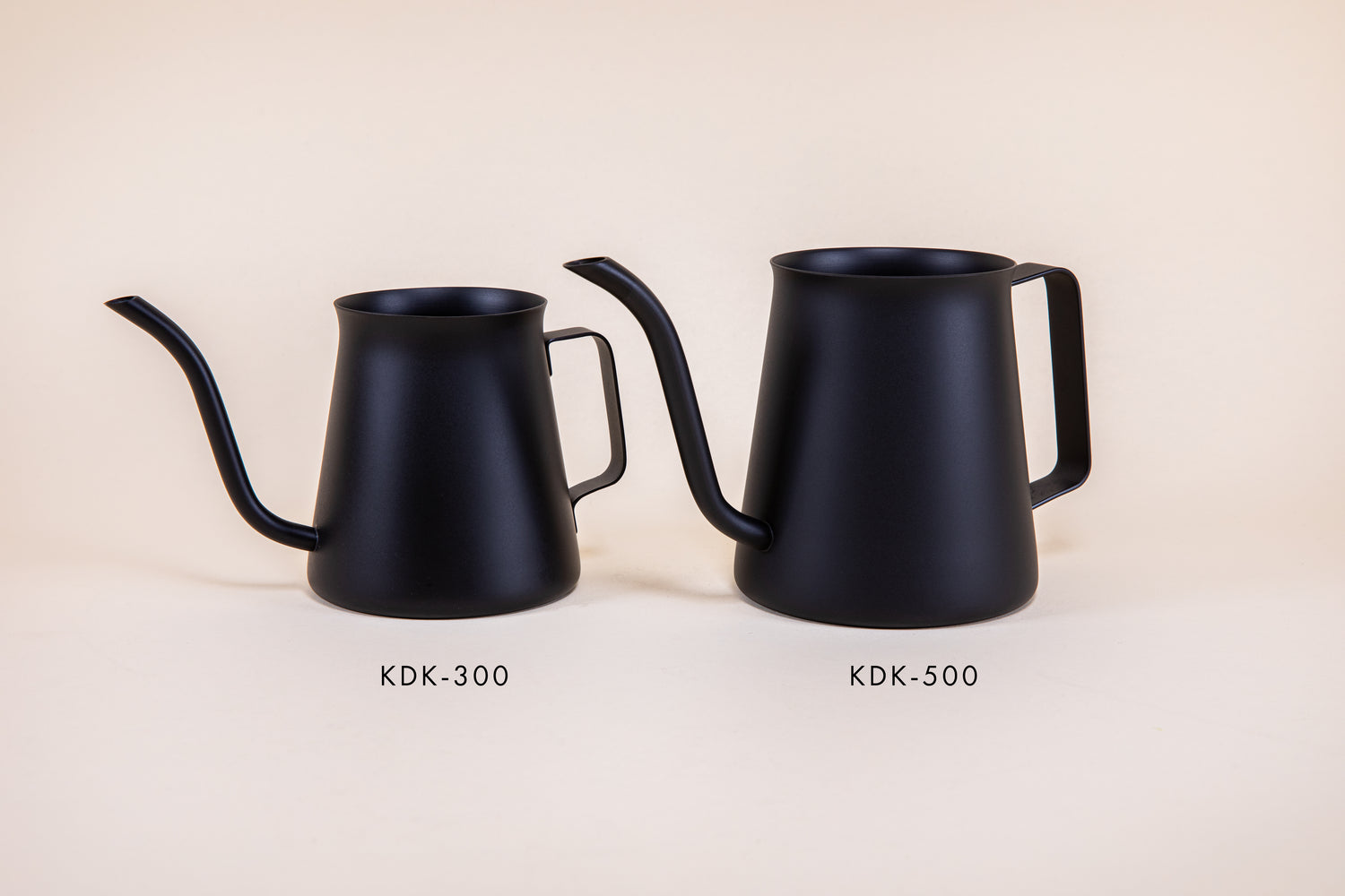 Small all black metal kettle with flared top, gooseneck spout, and handle next to a bigger all black metal kettle with flared top, gooseneck spout, and handle on a light background.