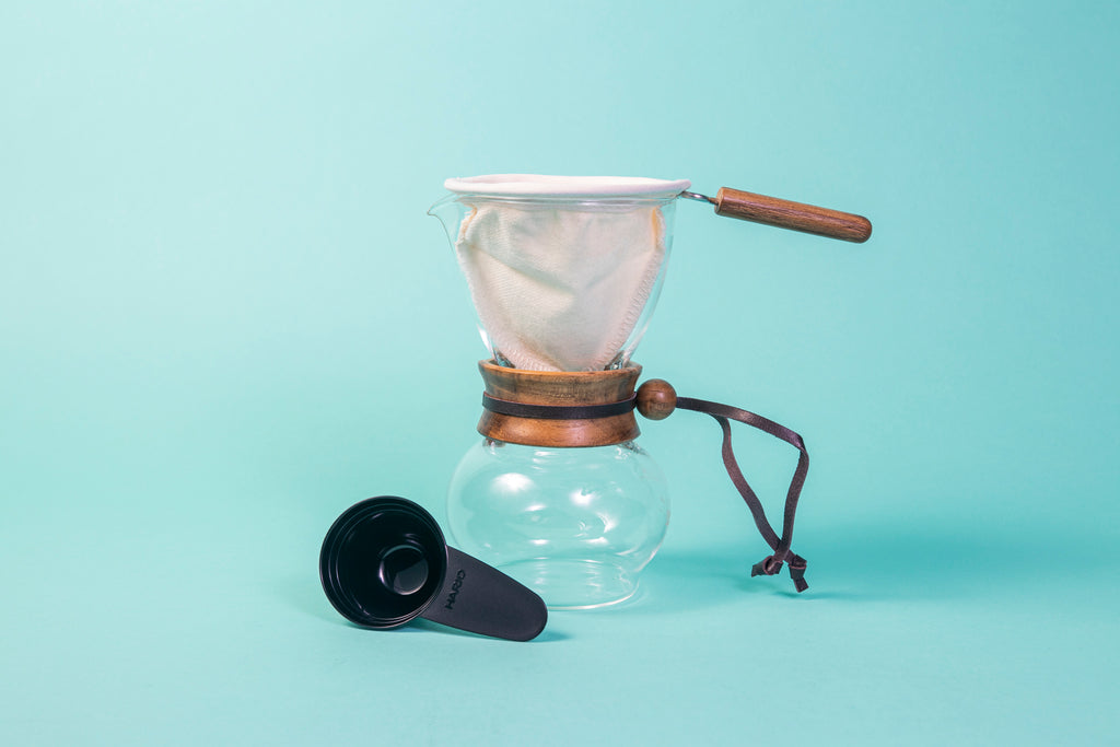 Glass server with a round base, acacia wood collar with leather tie, cloth filter and handle on top and a black plastic scoop.